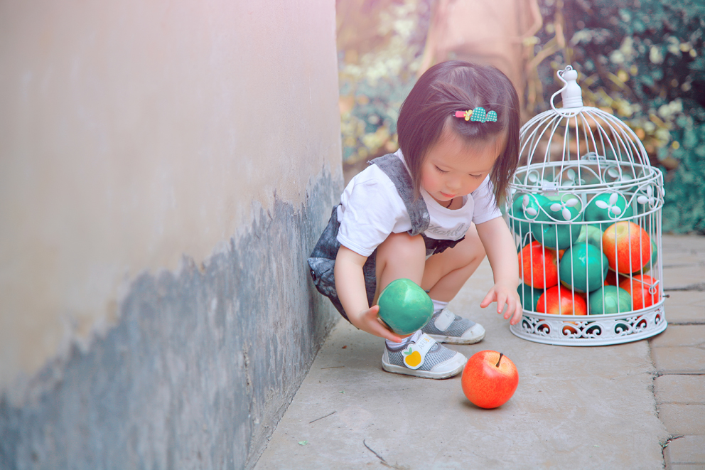 夏天应该就是色彩鲜艳的季节，小宝贝在红墙下独自嬉戏，红的苹果绿的苹果装满一个鸟笼，是的鸟笼，谁说装苹果一定要用水果篮啦，看这个小天使就在和笼子里的苹果玩的很开心。